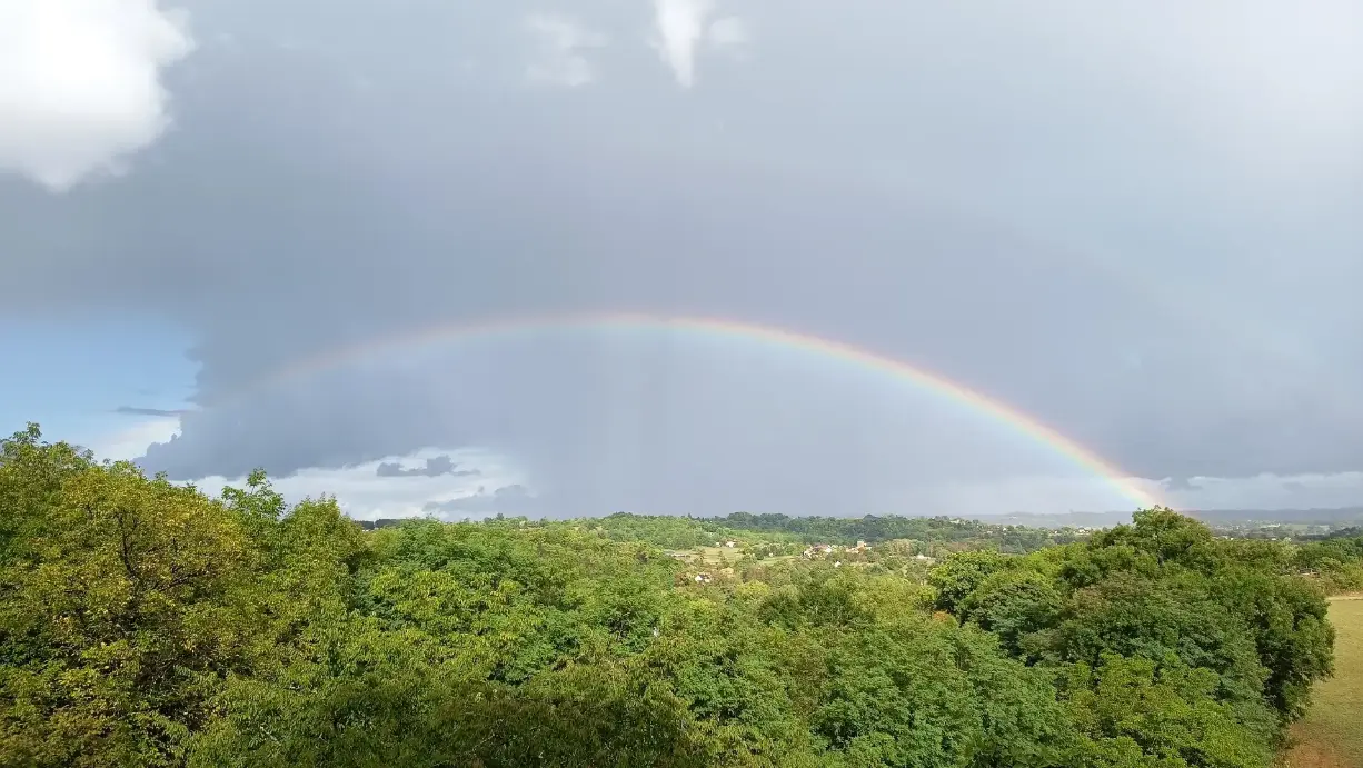 Arc-en-ciel complet et parfaitement arqué embrassant toute la vallée du Périgord Vert, vu depuis les hauteurs de La Fontaine du Mas, sur fond de ciel gris orageux et de forêts verdoyantes