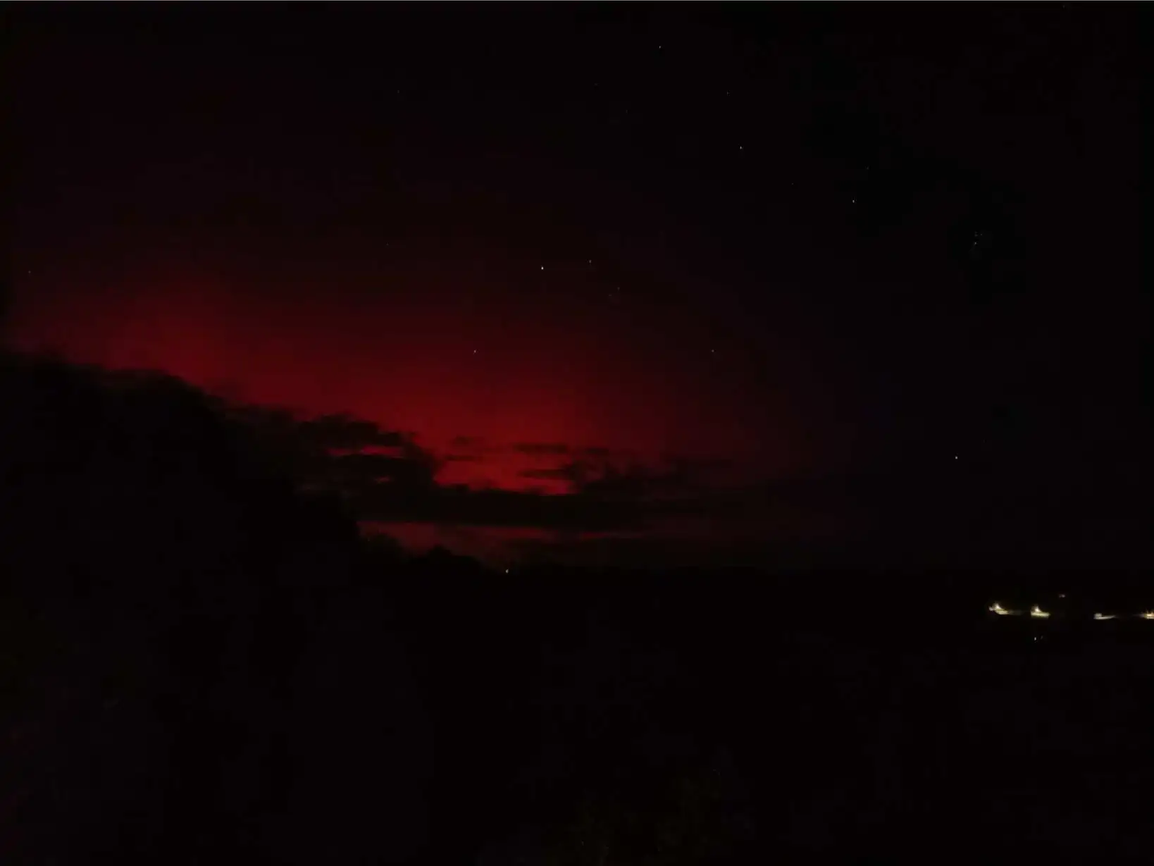 Aurore boréale rouge sang photographiée depuis La Fontaine du Mas : large lueur rouge cramoisie embrasant le ciel nocturne au-dessus de la ligne d'horizon sombre du Périgord, quelques étoiles visibles et lumières de villages au loin