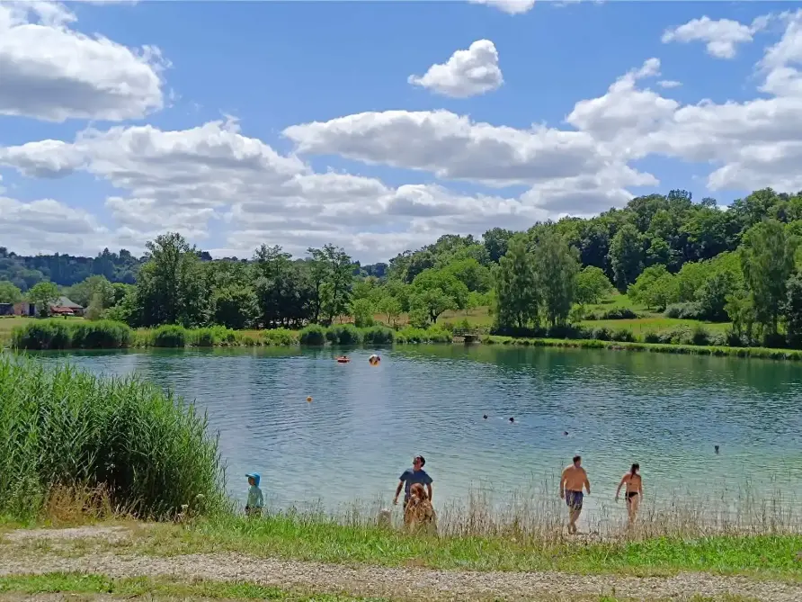 Baignade estivale au plan d'eau de Nantheuil : familles et enfants au bord du lac aux eaux turquoise entouré de roseaux et de forêts, sous un ciel bleu pomponné de nuages blancs