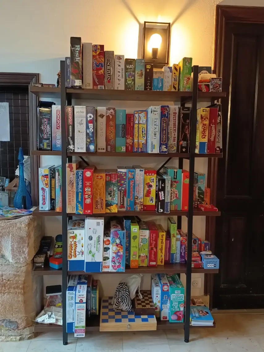 Grande bibliothèque en métal sombre remplie de jeux de société pour tous les âges à La Fontaine du Mas : Risk, Monopoly, Cluedo, Dixit, Scrabble, Puissance 4, Jungle Speed et dizaines d'autres, sur fond de mur en pierre ancienne