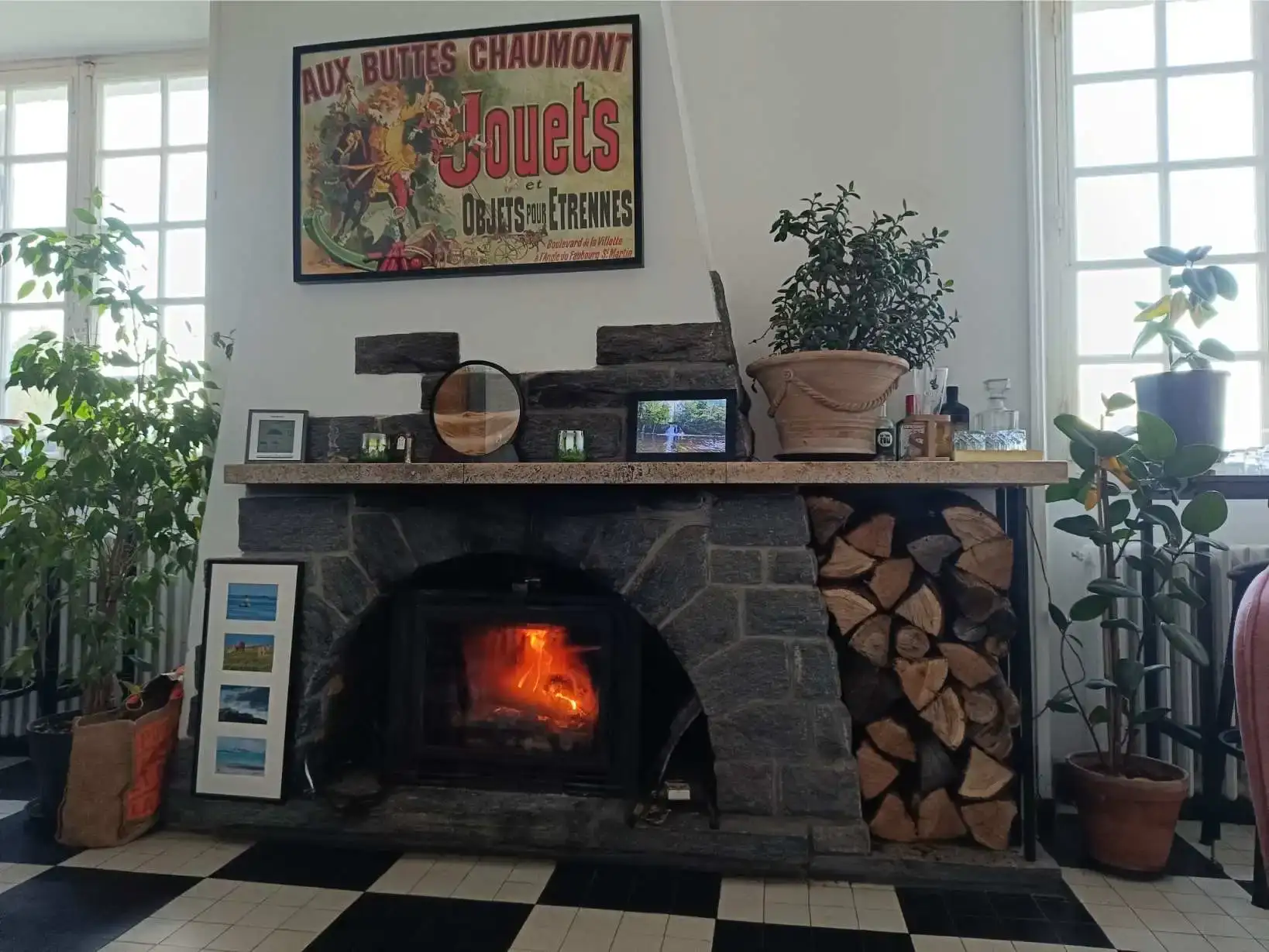 Grande cheminée en pierre grise du salon de La Fontaine du Mas avec feu vif allumé, rangée de bûches à droite, tablette de cheminée décorée d'objets et cadres photos, affiche ancienne au-dessus, grandes fenêtres à carreaux et plantes vertes de chaque côté