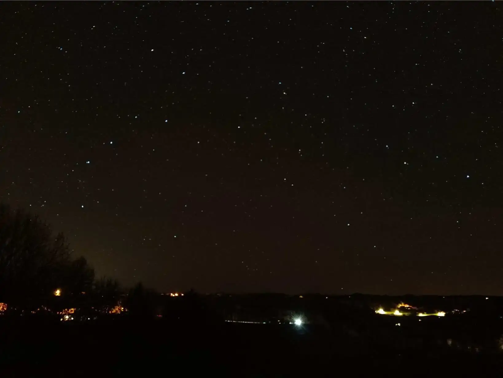 Ciel étoilé nocturne photographié en pose longue depuis La Fontaine du Mas avec des nuages fins traversant le champ d'étoiles, ligne d'horizon lumineuse des villages du Périgord Vert, végétation sombre au premier plan