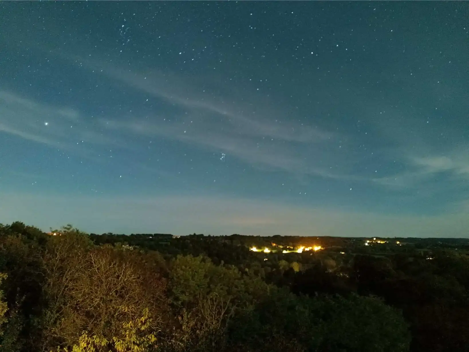 Ciel étoilé photographié en pose longue depuis La Fontaine du Mas : centaines d'étoiles visibles dans le ciel bleu nuit au-dessus de la vallée, lumières de Thiviers et des villages environnants dessinant la ligne d'horizon, preuve de la faible pollution lumineuse du Périgord Vert