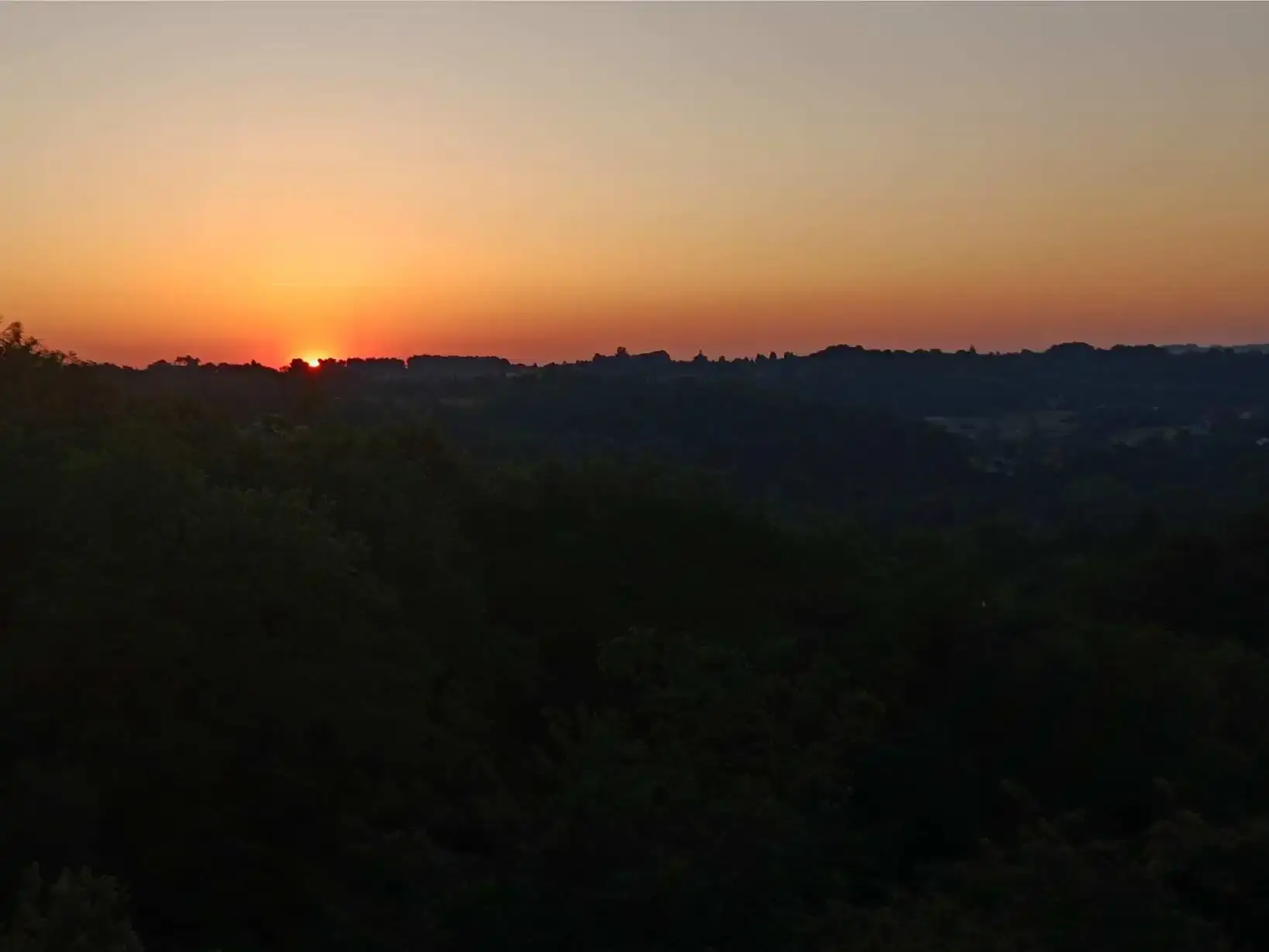 Coucher de soleil sur les collines boisées du Périgord Vert depuis La Fontaine du Mas : dernière lueur orangée à l'horizon se fondant dans un ciel dégradé du doré au mauve, silhouettes des collines et de la végétation au premier plan