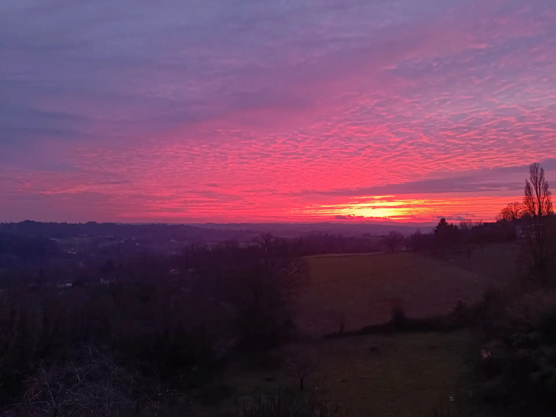 Coucher de soleil ardent sur la vallée du Périgord depuis La Fontaine du Mas : disque solaire orange-rouge posé sur la ligne de crête boisée sombre, ciel dégradé du rouge vif à l'or pâle, silhouettes d'arbres et toits de Thiviers