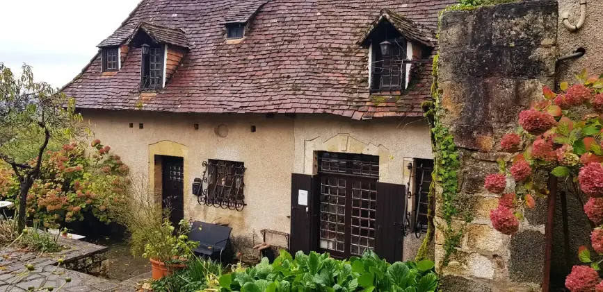 Façade de la dépendance ancienne de La Fontaine du Mas en automne : toits de lauzes brunes, volets et fenêtres à petits carreaux en fer forgé, hortensias roses tardifs et chou vert au premier plan
