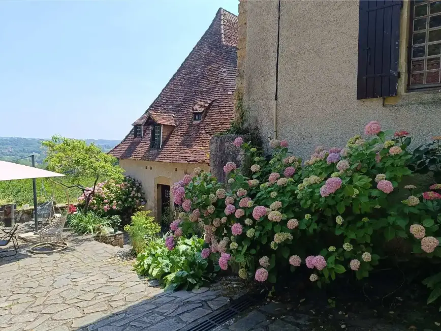 Façade de La Fontaine du Mas en été entourée d'hortensias roses et blancs en pleine floraison, toits de tuiles ocre, terrasse dallée avec table et chaise suspendue visibles, vue dégagée sur la vallée boisée en arrière-plan