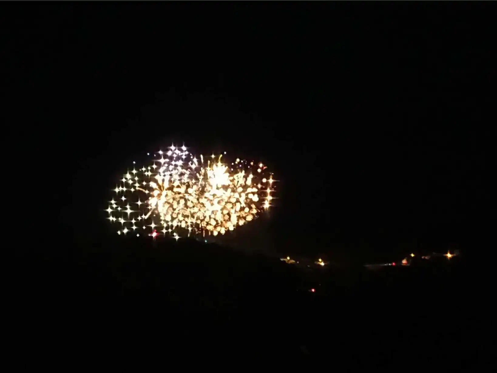 Feu d'artifice blanc et doré éclatant dans le ciel nocturne au-dessus de la vallée du Périgord, visible depuis les hauteurs de La Fontaine du Mas, avec quelques lumières de villages en contrebas