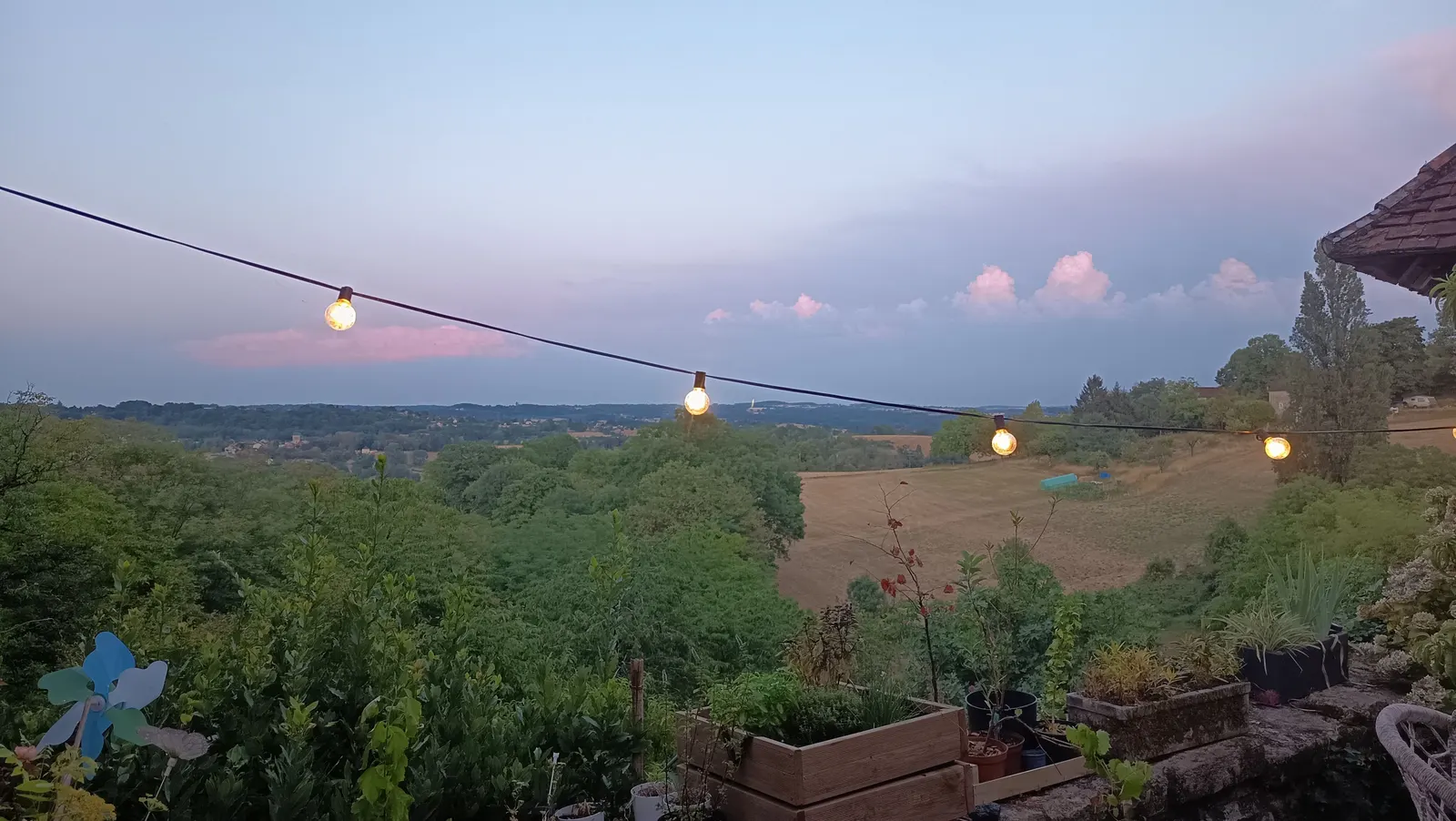 Guirlande guinguette allumée au crépuscule, tendue au-dessus du potager en bacs, avec vue sur la vallée du Périgord et un ciel rosé strié de nuages