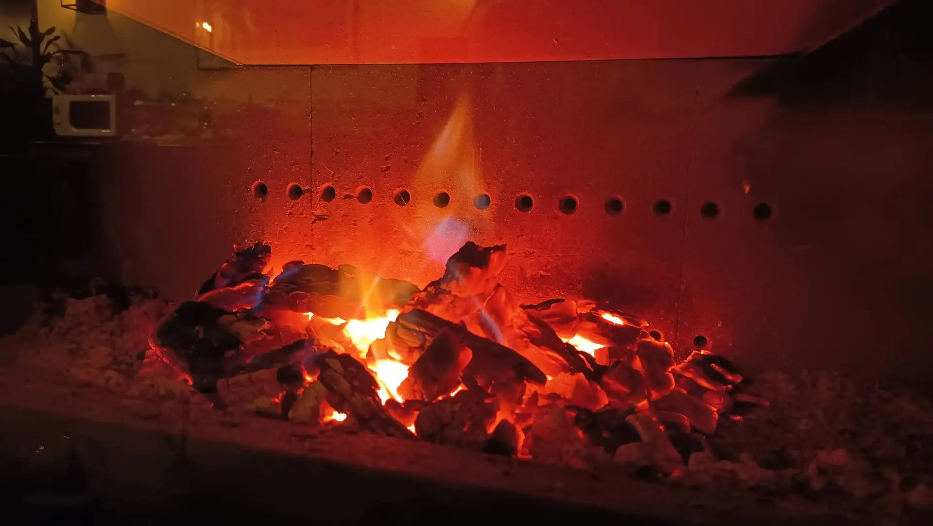 Insert de cheminée à braises rougeoyantes dans le salon de La Fontaine du Mas : bûches incandescentes et braises orangées dans un foyer en béton réfractaire perforé de trous d'aération, unique flamme bleue et jaune au centre, lueur rouge sang enveloppant toute la chambre de chauffe