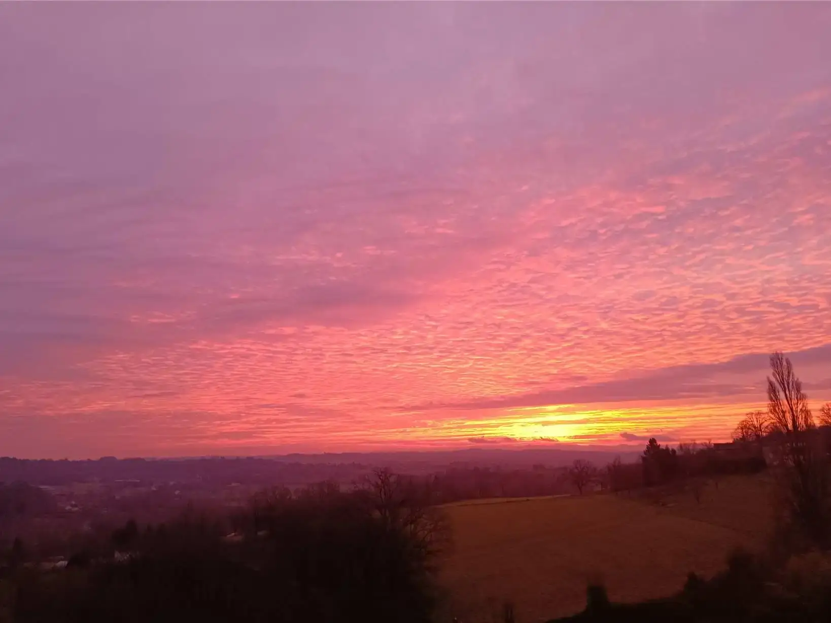 Lever de soleil spectaculaire en hiver depuis La Fontaine du Mas : ciel entièrement embrasé de roses, mauves et orangés vifs sur un fond de nuages en écailles, prairie dorée et silhouettes de collines du Périgord, lumière rasante irréelle