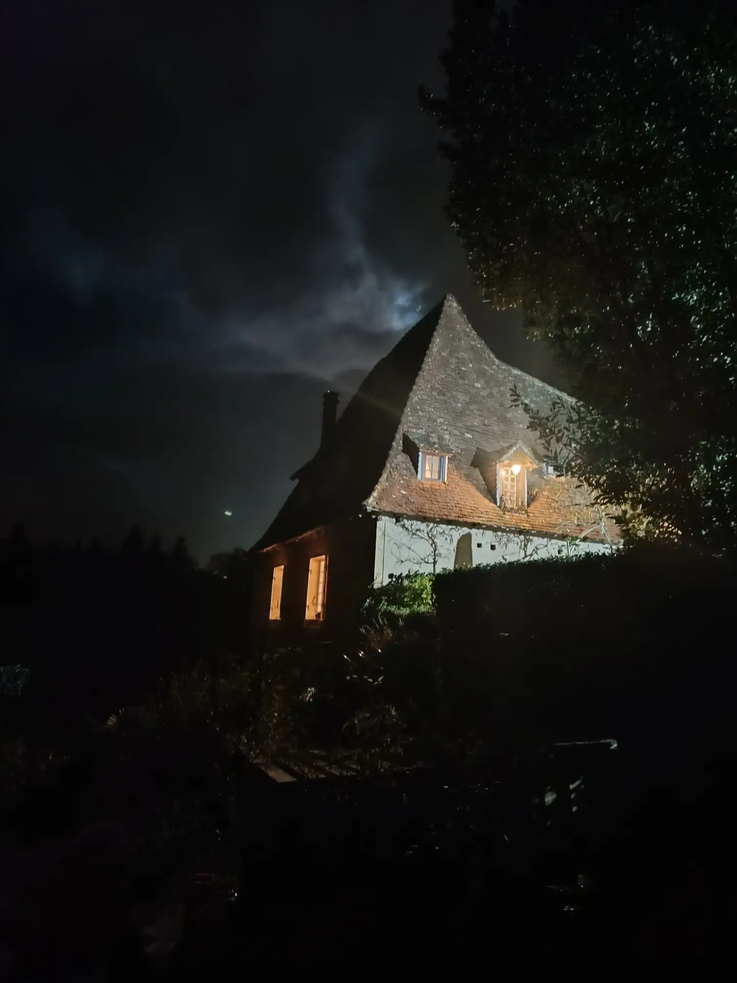 La Fontaine du Mas photographiée de nuit : toiture pointue en lauzes de la dépendance se détachant sur un ciel nocturne nuageux, fenêtres illuminées d'une lumière chaude dorée, arbres et haies en ombre, atmosphère intimiste et mystérieuse