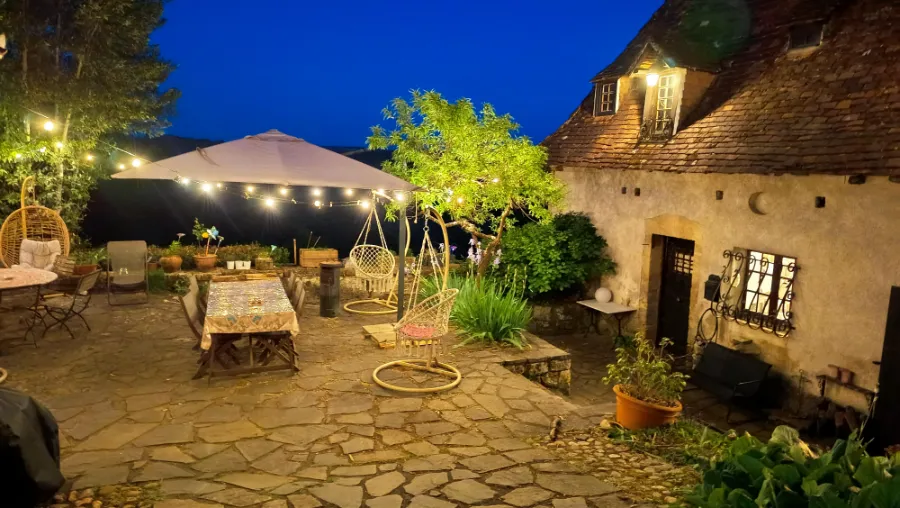 Stone terrace at La Fontaine du Mas lit up with fairy lights at dusk, with garden table, rattan hanging chairs and white parasol in front of the old stone house