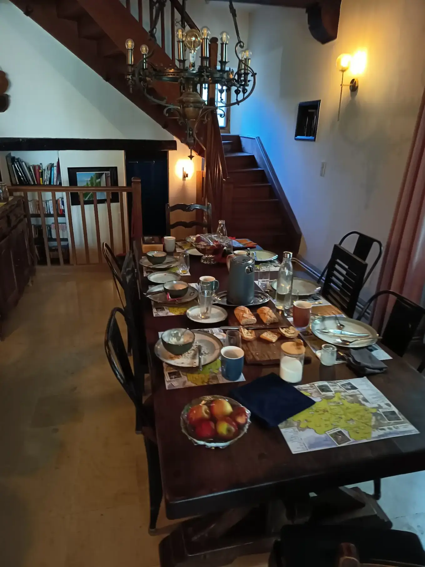 Table du petit-déjeuner en cours de repas dans la salle à manger de La Fontaine du Mas : cafetière, pain tranché, pommes, cartes touristiques, tasses et vaisselle sur la grande table en bois sombre, escalier en bois et lustre en fer forgé en arrière-plan