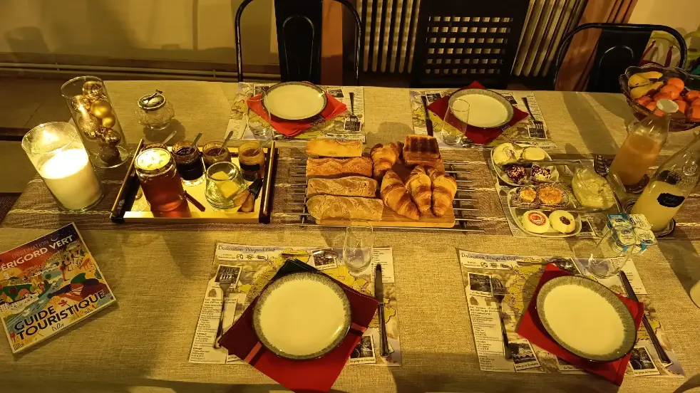 Table du petit-déjeuner de Noël dressée à La Fontaine du Mas : plateau de croissants, pain, viennoiseries, confitures, miel, beurre, jus d'orange et yaourts sur nappe beige, guide touristique Périgord Vert et bougeoirs décoratifs