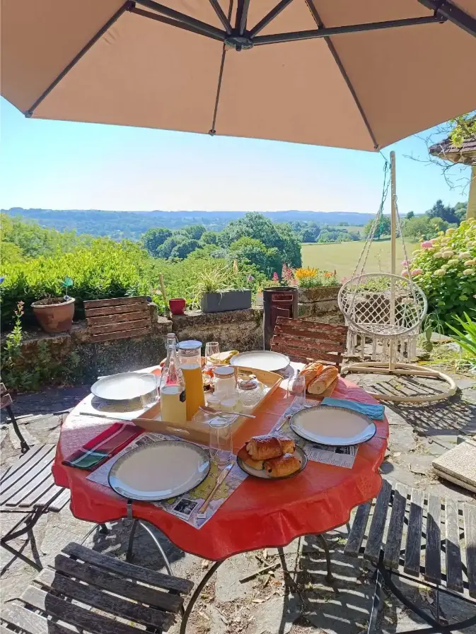 Petit-déjeuner servi en plein air sur la terrasse de La Fontaine du Mas : table ronde nappe rouge avec croissants, baguette, jus d'orange et confitures sous un parasol beige, vue panoramique sur la campagne verdoyante et chaise suspendue en macramé