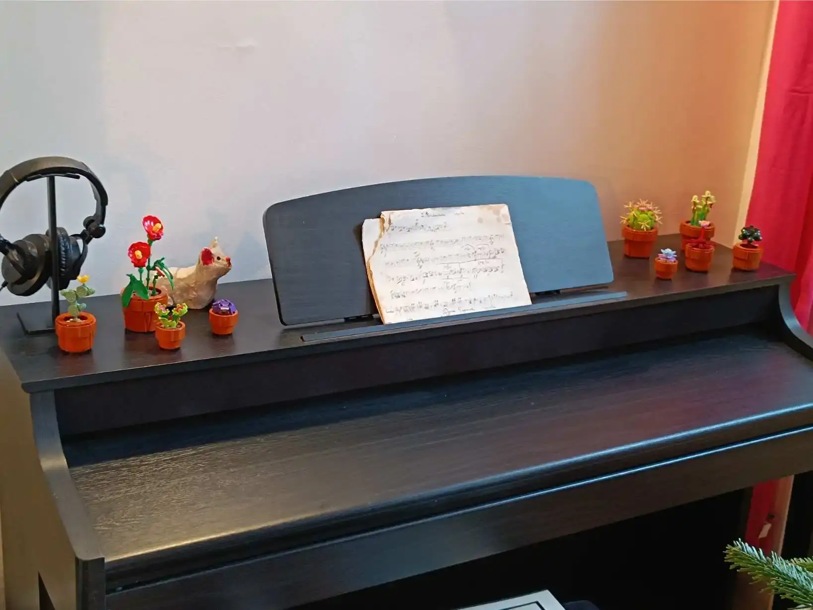 Piano numérique noir avec partition ancienne usée posée sur le pupitre, petites plantes en pots décoratifs Lego et figurine sur le dessus, casque audio sur support à gauche, décoration de Noël visible en bas à droite, La Fontaine du Mas