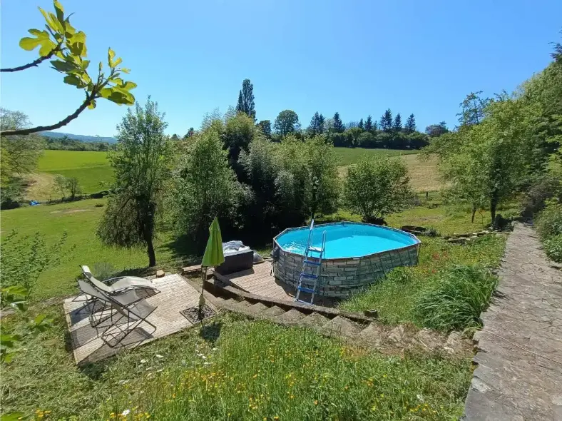 Piscine hors-sol ronde de La Fontaine du Mas avec terrasse en bois, transat et parasol vert, entourée d'herbe fleurie et de pissenlits, avec vue ouverte sur la campagne du Périgord Vert