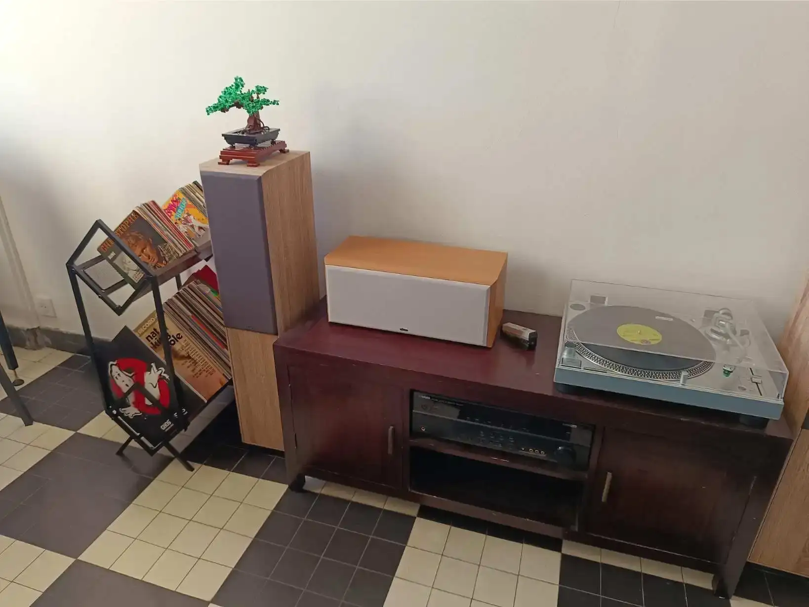 Platine vinyle avec disque jaune posé sur un meuble rouge sombre, enceinte centrale en bois, collection de vinyles dans un présentoir noir à gauche avec pochettes colorées dont Ghostbusters visible, bonsaï décoratif Lego sur la colonne d'enceinte, sol en damier noir et blanc