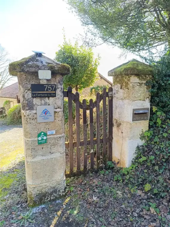 Portail d'entrée de La Fontaine du Mas : deux piliers en pierres de taille moussues avec plaque numéro 757, enseigne La Fontaine du Mas, label Chambres d'Hôtes et badge Accueil Vélo, petit portillon en bois peint en brun