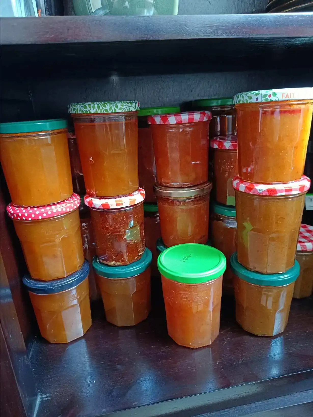 Rangée de pots et bocaux de confiture maison à la couleur orange ambrée, couvercles variés — rouge à pois, vichy rouge, vert —, alignés sur une étagère en bois sombre, production artisanale de La Fontaine du Mas