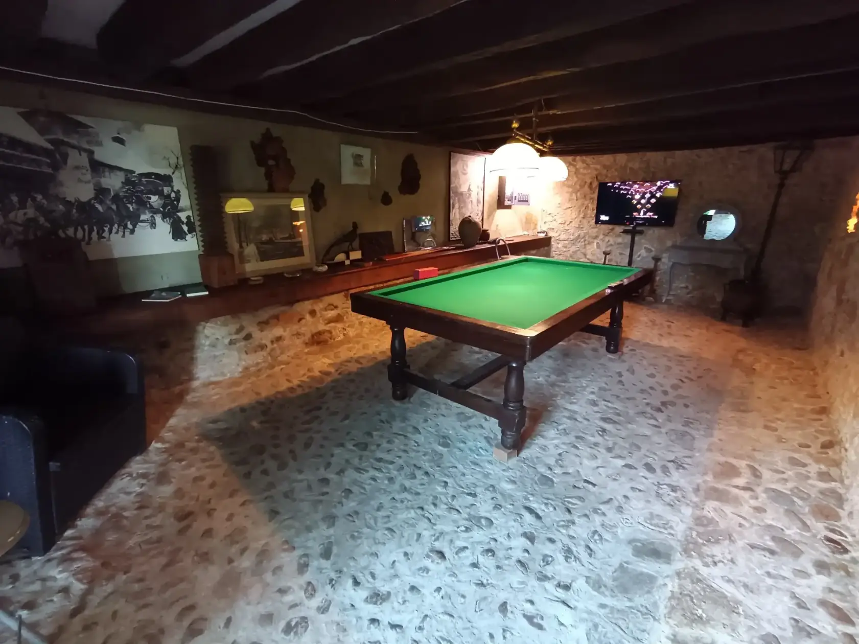 Salle de jeux de La Fontaine du Mas avec billard français sur tapis vert, murs en pierres apparentes, télévision murale et fauteuil en cuir noir, éclairage tamisé