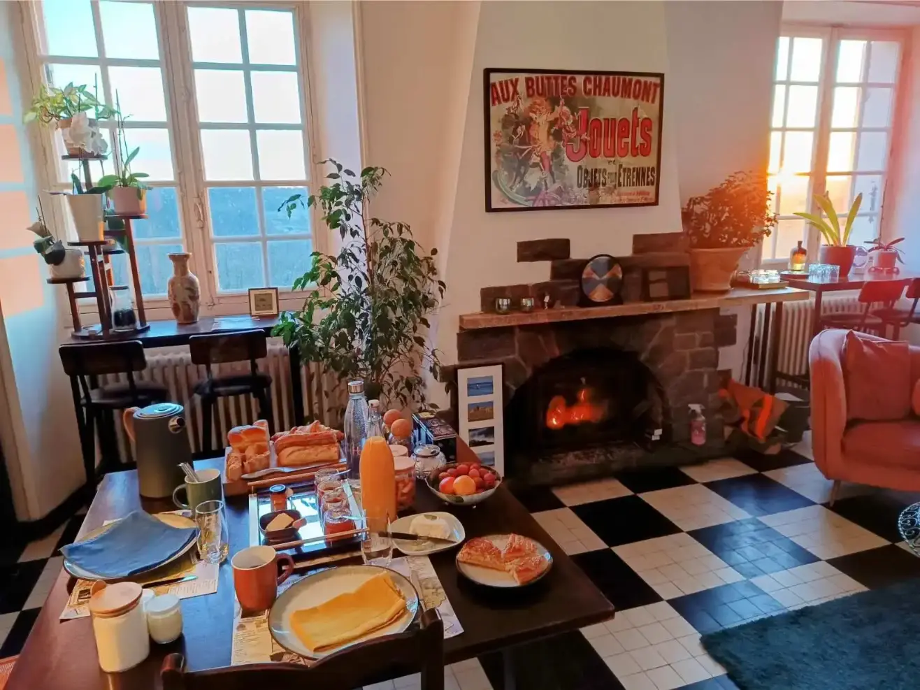 Salon de La Fontaine du Mas un matin d'automne : table de petit-déjeuner avec pain, jus d'orange et fruits, devant une cheminée en briques avec feu allumé, sol en damier noir et blanc, affiche ancienne et grande fenêtre à carreaux donnant sur la campagne