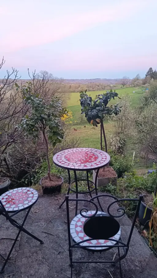 Salon de jardin en fer forgé mosaïque rouge et blanc sur la terrasse de La Fontaine du Mas au crépuscule printanier, avec vue sur la prairie et les ânes visibles en contrebas dans les dernières lueurs roses du soir