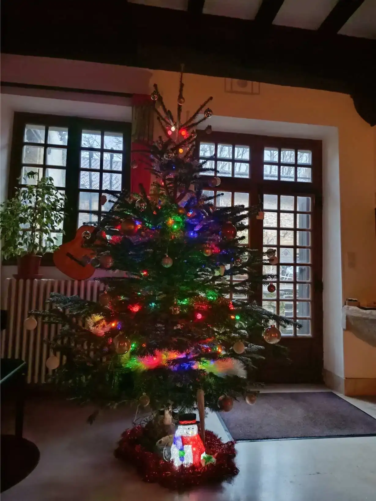 Sapin de Noël décoré avec guirlandes lumineuses multicolores, boules et figurine de bonhomme de neige lumineux à la base, dans le salon de La Fontaine du Mas avec grandes fenêtres à carreaux et poutres apparentes au plafond, guitare acoustique rouge visible en arrière-plan