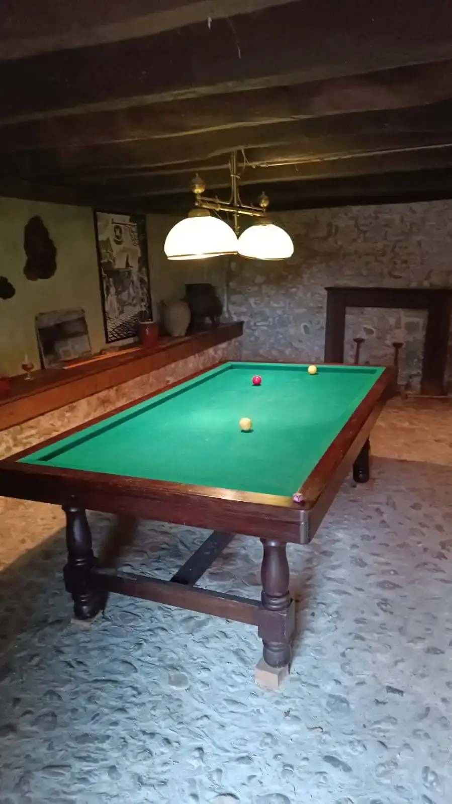Table de billard française en bois sombre sur tapis vert avec trois billes (rouge, blanche et jaune) dans la salle de jeux voûtée de La Fontaine du Mas : murs en pierre de taille apparente, plafond à poutres sombres, lustre en laiton doré à trois globes blancs, cheminée ancienne en pierre et sol en galets ronds