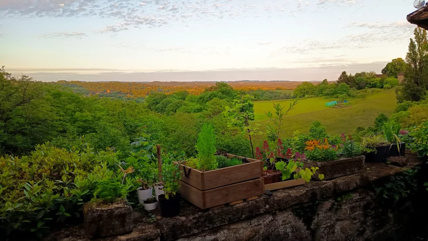 Terrasse en pierre de La Fontaine du Mas au coucher du soleil, avec jardinière en bois et pots de fleurs colorés au premier plan, surplombant un panorama sur la campagne verdoyante du Périgord Vert