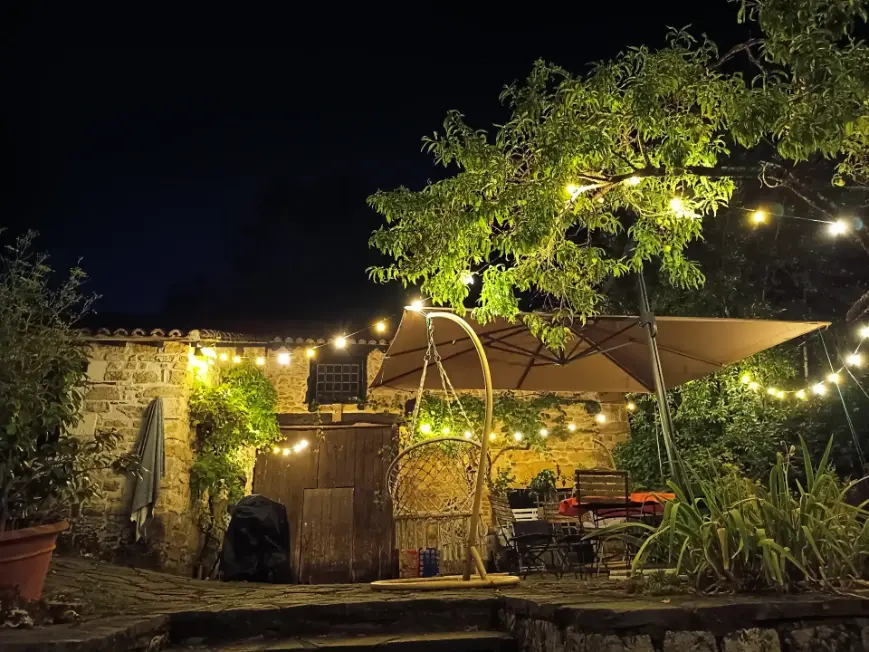 Terrasse nocturne de La Fontaine du Mas illuminée par des guirlandes lumineuses chaleureuses accrochées entre les arbres et la façade en pierre, avec chaise suspendue en macramé, parasol et salon de jardin dans la pénombre