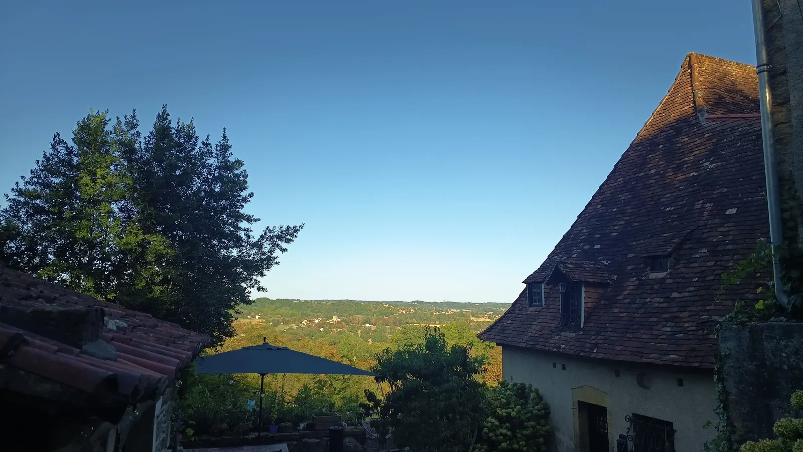 Toits de tuiles brunes du bâtiment principal se découpant sur un ciel bleu d'été, avec vue dégagée sur la vallée boisée en contrebas