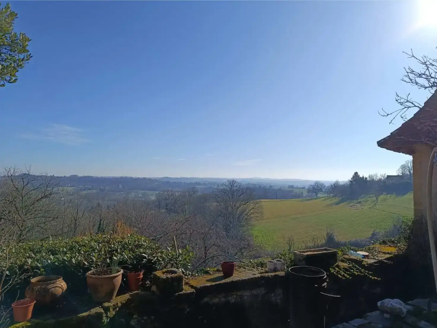Vue panoramique depuis le muret du jardin de La Fontaine du Mas sur la campagne du Périgord Vert, pots de fleurs au premier plan, prairies et bois à perte de vue sous un ciel dégagé