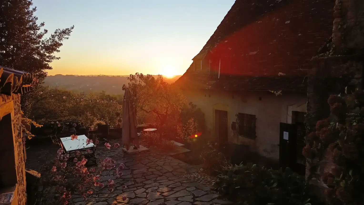Vue depuis le toit de La Fontaine du Mas sur la terrasse au coucher du soleil, avec parasol et mobilier de jardin dans la lumière dorée du soir