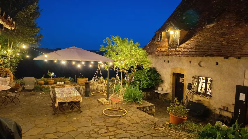 Terrasse en pierre de La Fontaine du Mas illuminée en soirée, avec guirlandes lumineuses, parasol et table dressée dans la cour intérieure du manoir périgordin
